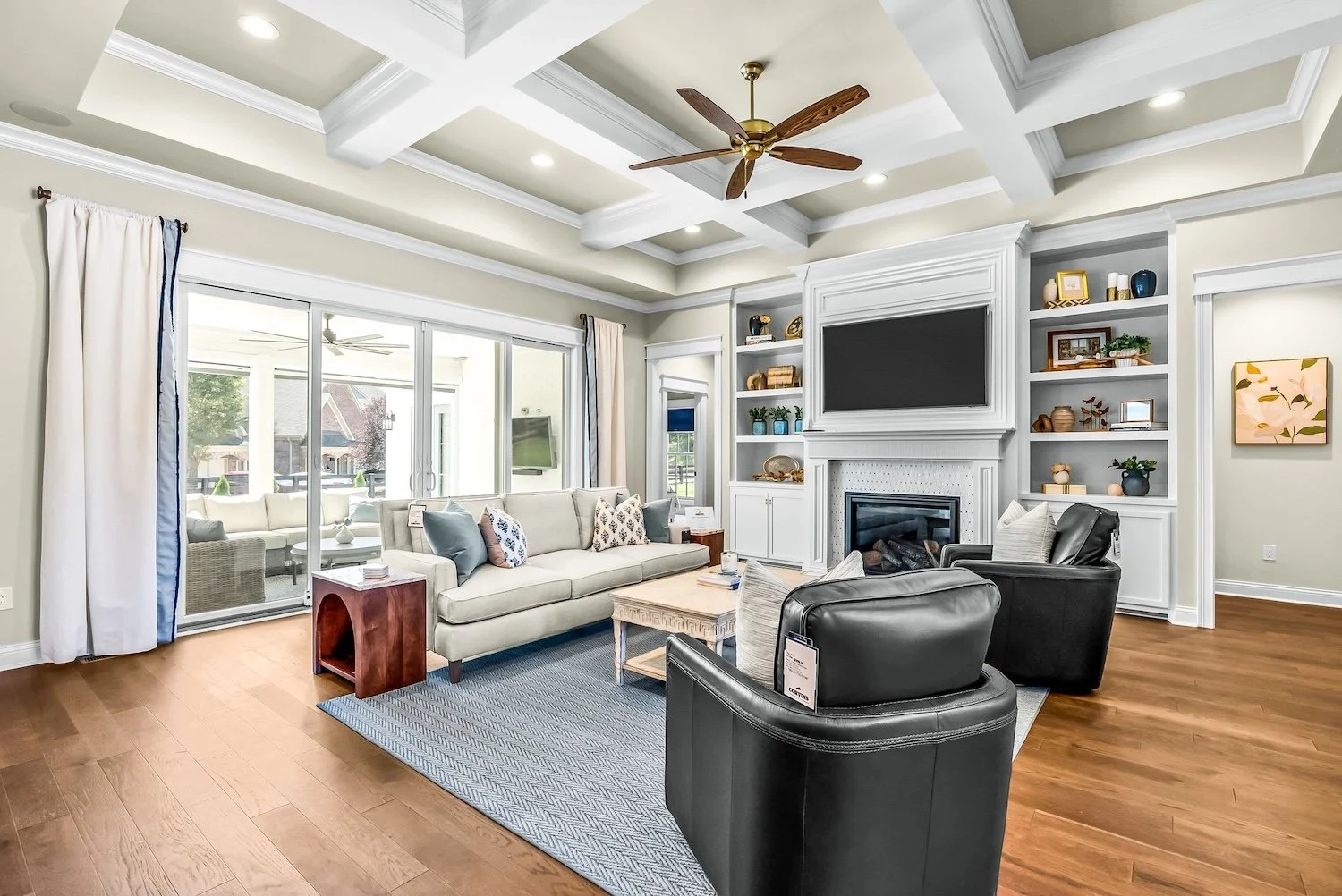 Living room with beige walls, a white coffered ceiling, and a wooden floor. It has a white sofa with decorative pillows, two black leather armchairs, a wooden coffee table, and a blue area rug. There is a white fireplace with built-in shelves and a l