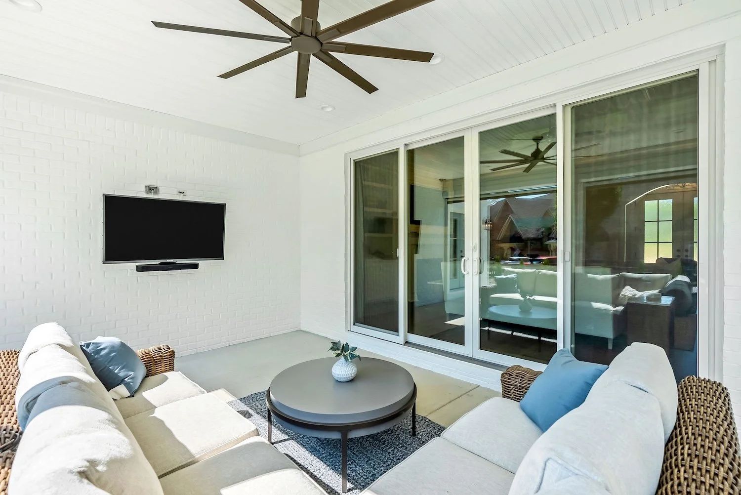 Covered outdoor patio with white brick walls, a ceiling fan, a wall-mounted flat-screen TV, a round coffee table with a small potted plant, and outdoor sofas with cushions, overlooking the street through sliding glass doors.