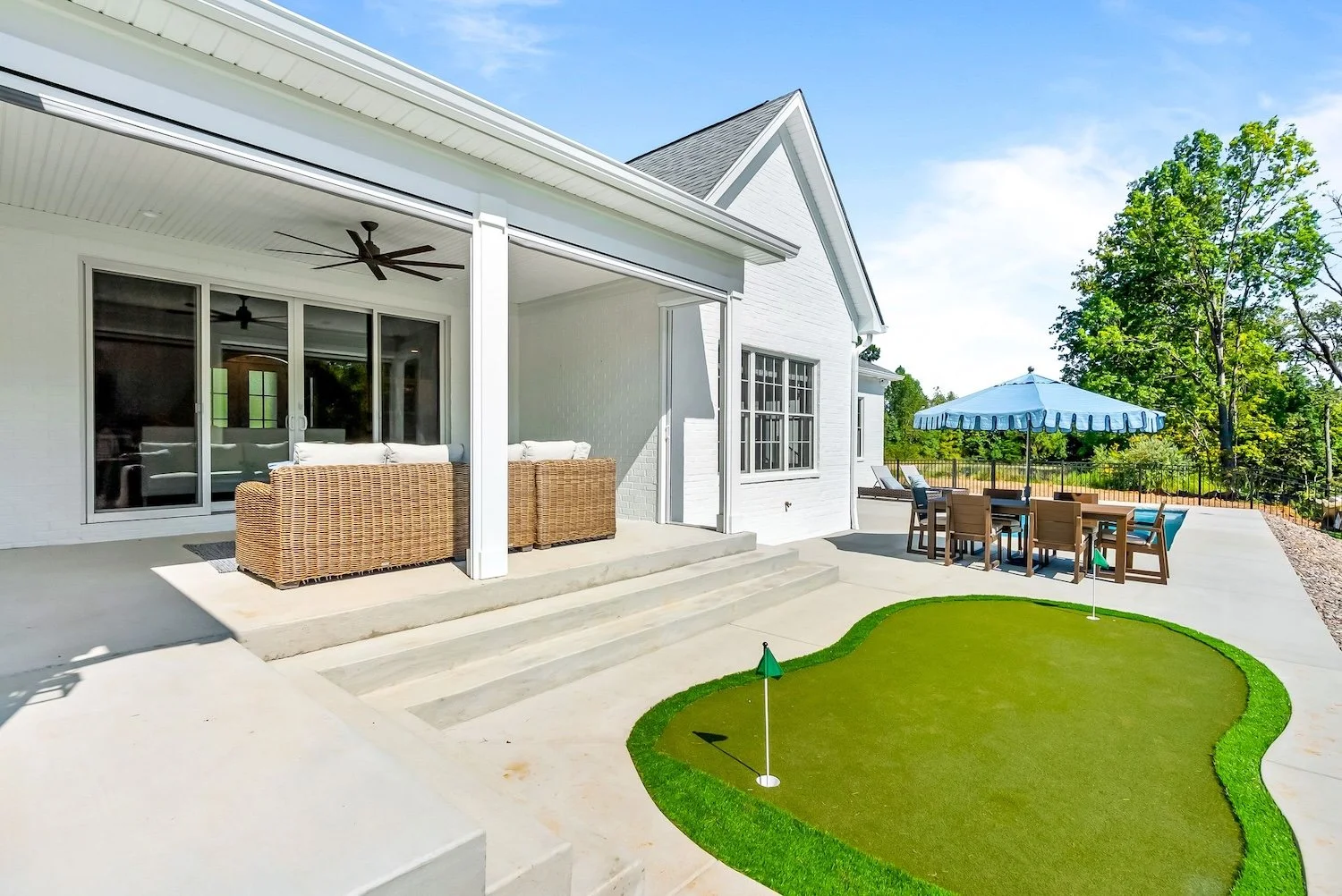 A backyard patio with a covered seating area, outdoor dining table with chairs under a large umbrella, and a mini golf putting green, with trees and a fence in the background.