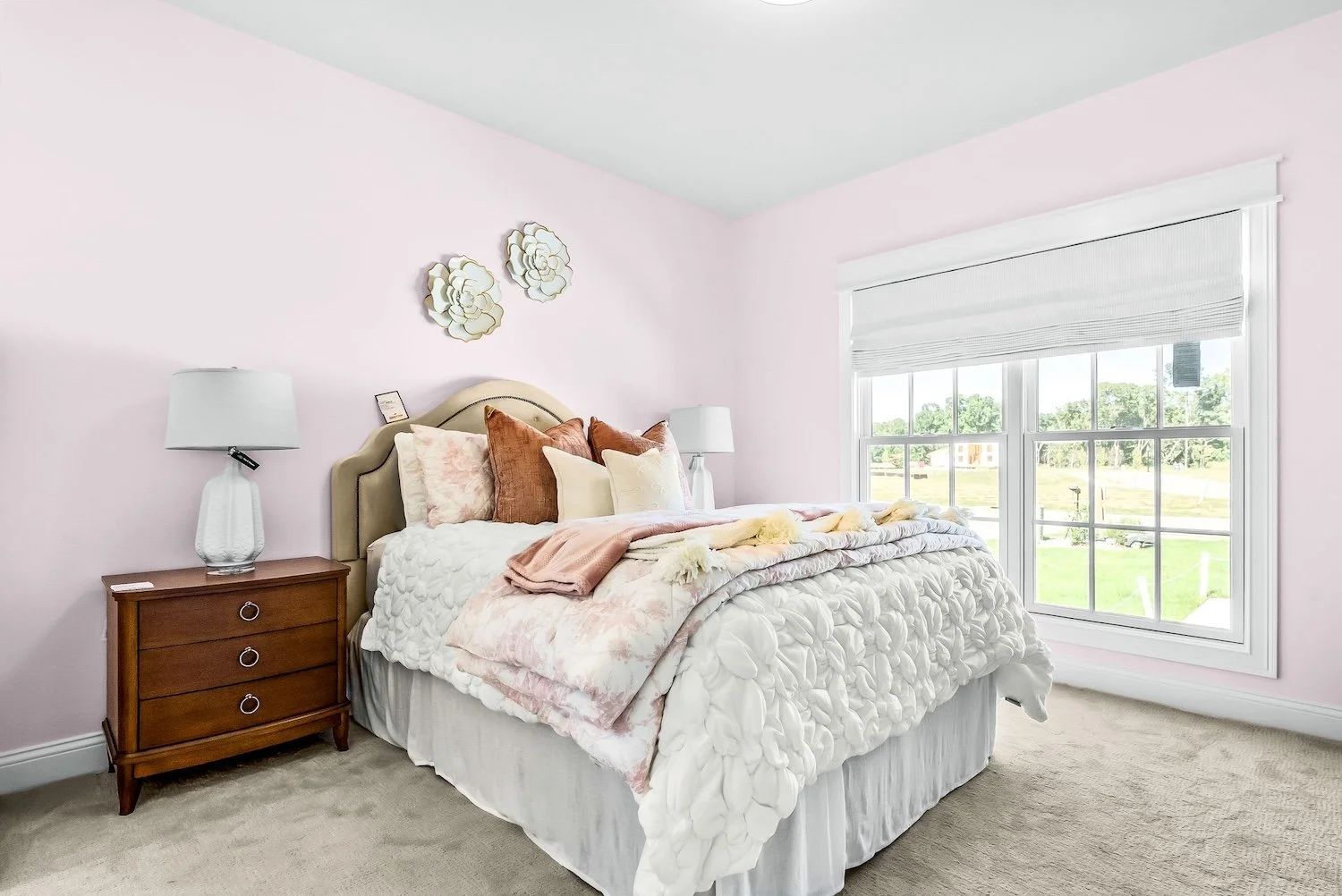 A bedroom with light pink walls, a large window with white blinds, a beige upholstered headboard, a white bedspread with floral textures, multiple pillows including white, beige, and rust-colored ones, a pink blanket, and a wooden nightstand with a w