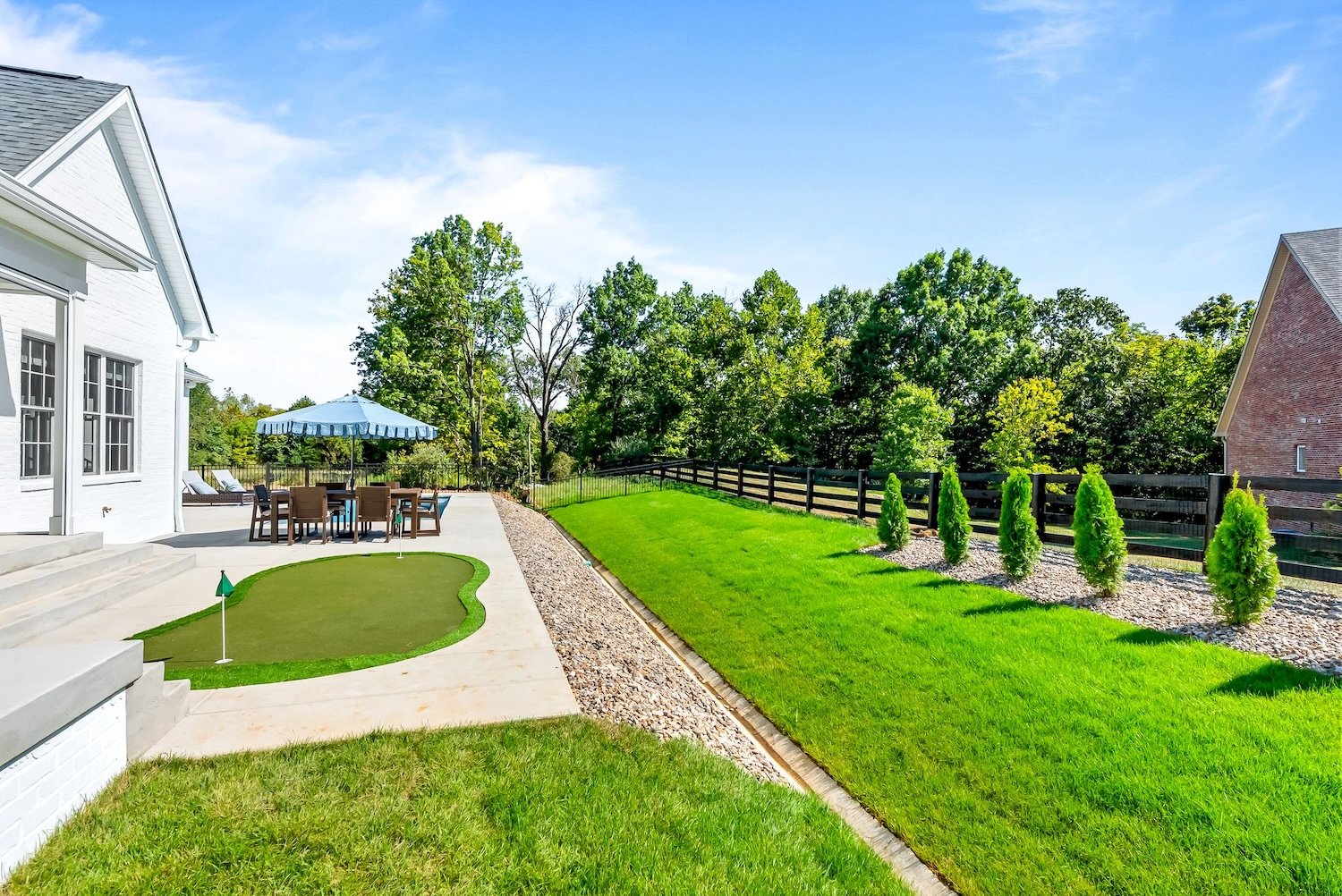 Backyard with green grass, a patio area with a table and chairs, a large blue umbrella, a small putting green, and a black wooden fence with small trees along it, surrounded by trees under a clear blue sky.