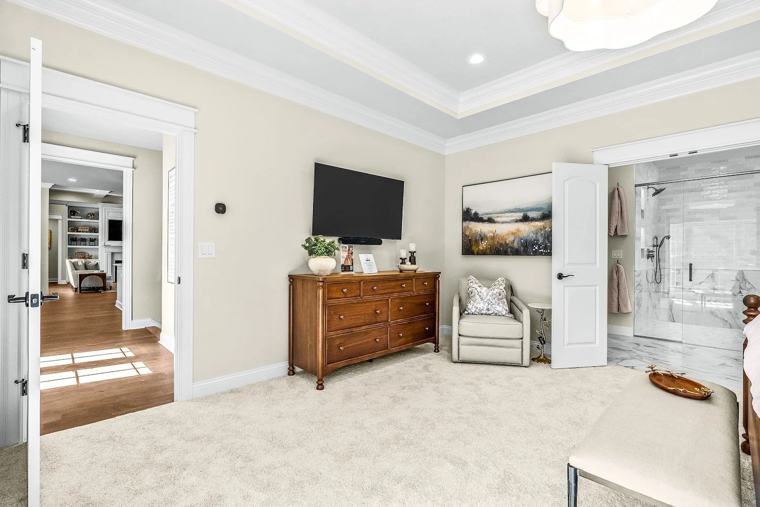 A bedroom with a doorway to a bathroom, a beige carpeted floor, a wall-mounted TV above a wooden dresser, a cream armchair with a decorative pillow, a landscape painting on the wall, and a glass shower enclosure with marbled tiles inside the bathroom