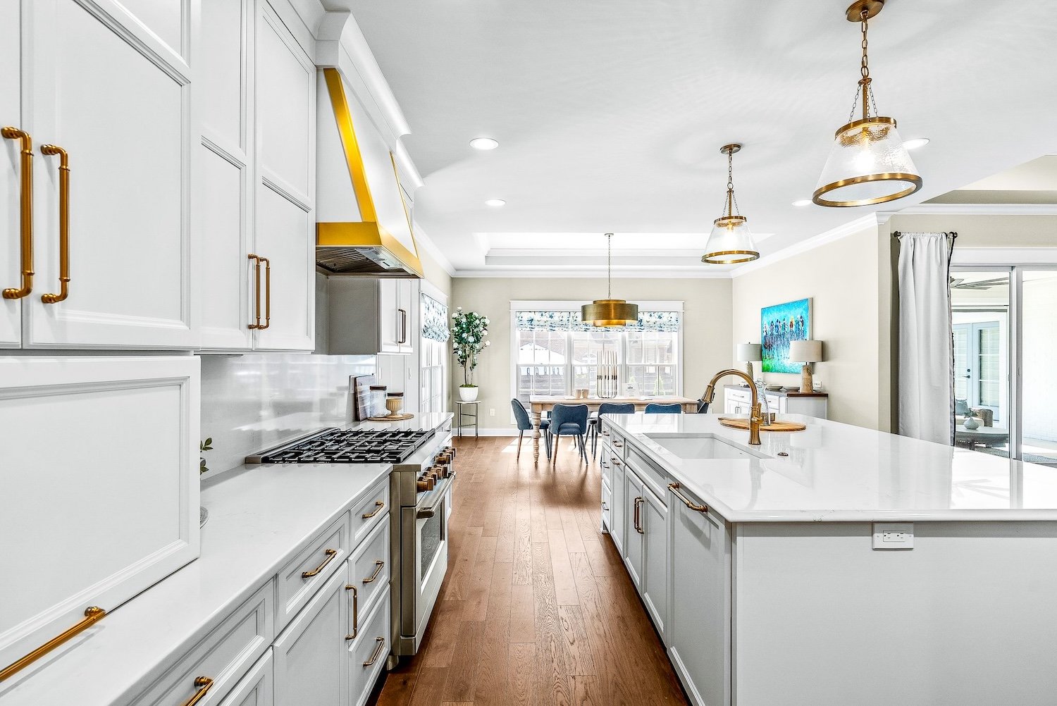 Bright, modern kitchen with white cabinetry, gold handles, a gold range hood, and a central island with a white countertop. Wooden floors, gold and glass pendant lights, a dining area with a table and blue chairs, and a living space with a wall-mount