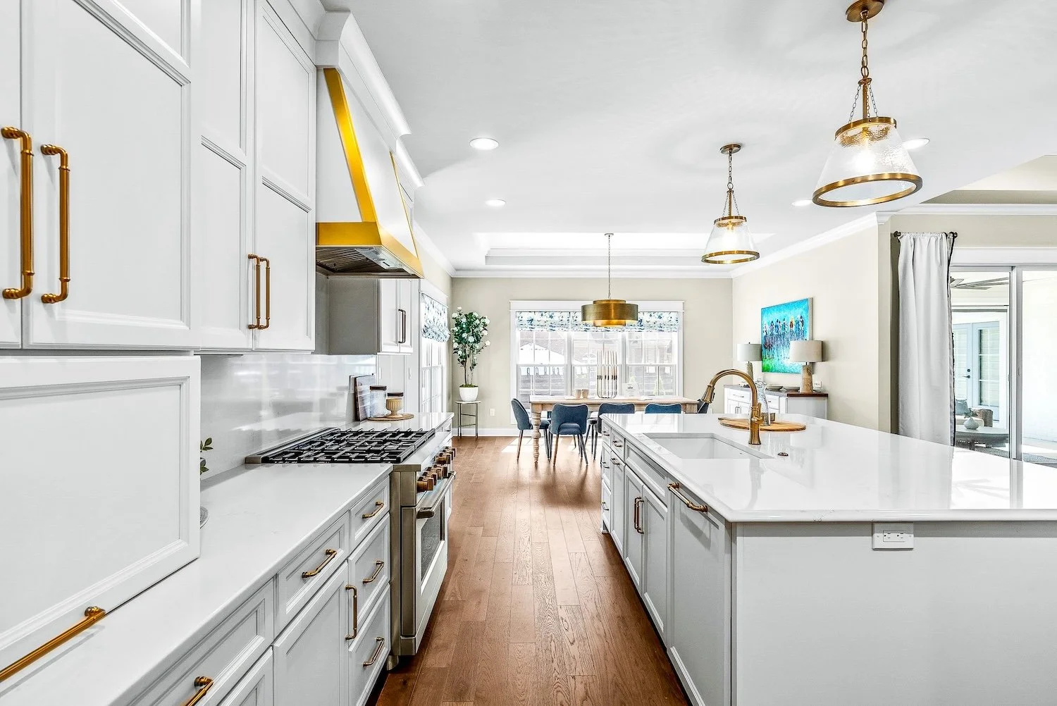 Bright, modern kitchen with white cabinetry, gold handles, a gold range hood, and a central island with a white countertop. Wooden floors, gold and glass pendant lights, a dining area with a table and blue chairs, and a living space with a wall-mount