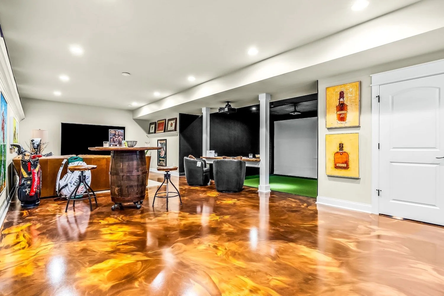 An indoor recreational room with polished, shiny wood floors, a bar area with chairs, a golf bag with clubs, a television, seating area with chairs, and wall art featuring bottle images. There is a section with a projector screen and small green turf
