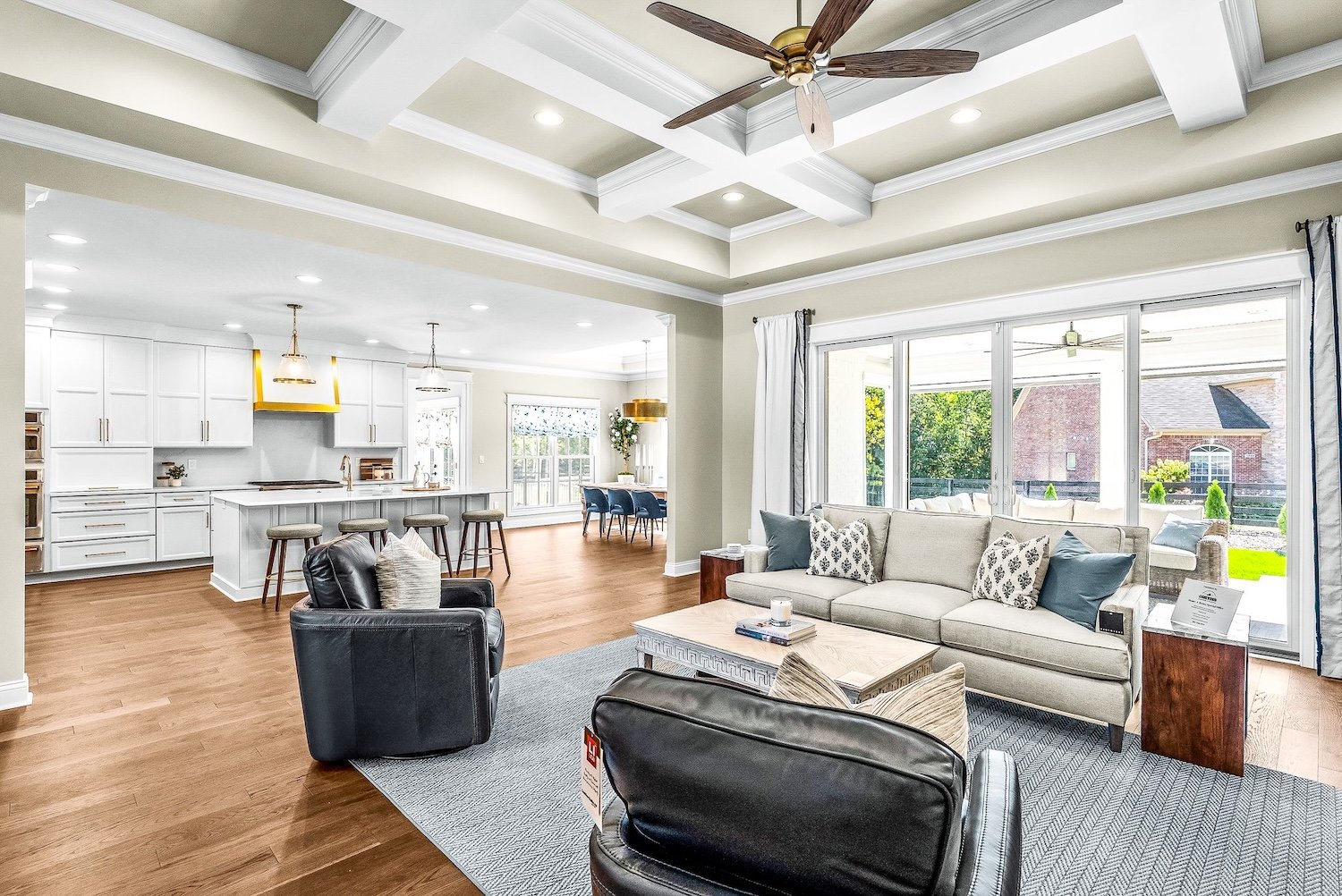 Open-concept living room and kitchen with hardwood floors, white cabinets, a kitchen island with bar stools, large windows, cream sofa, black leather armchairs, and a ceiling fan.