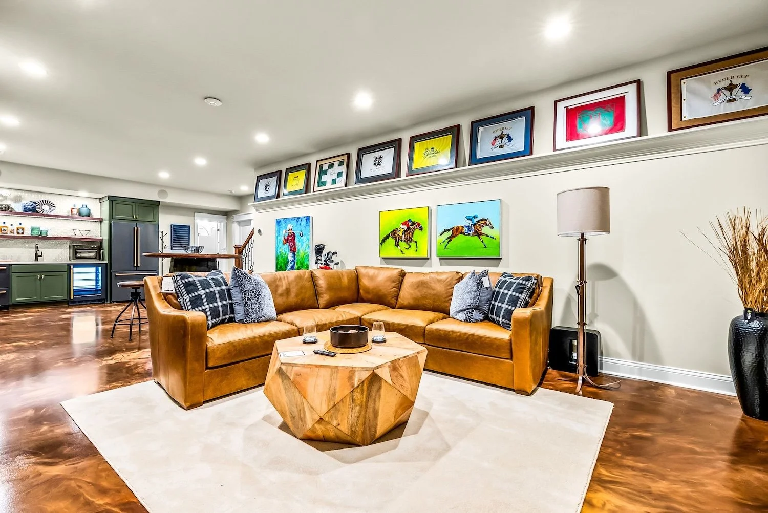 Living room with a tan leather sectional sofa, a wooden coffee table, and a white rug. The wall behind the sofa features colorful artwork and framed sports memorabilia. There is a tall floor lamp with a beige shade and a black vase with decorative dr