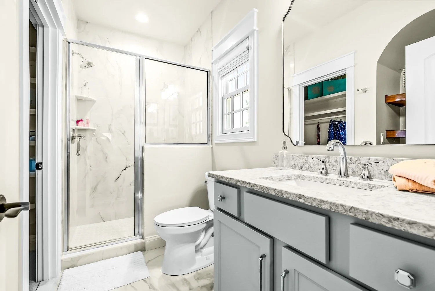 Bathroom with a walk-in shower with marble walls, a toilet, a gray vanity with a marble countertop, a mirror, and a window.
