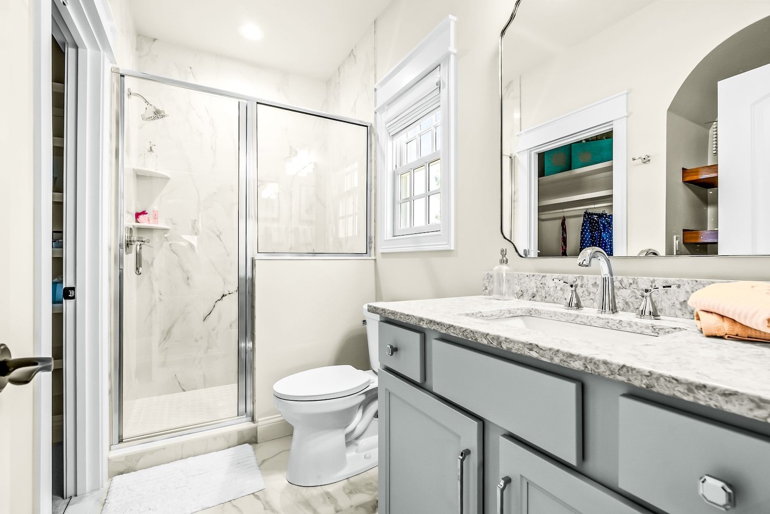 Bathroom with a walk-in shower with marble walls, a toilet, a gray vanity with a marble countertop, a mirror, and a window.