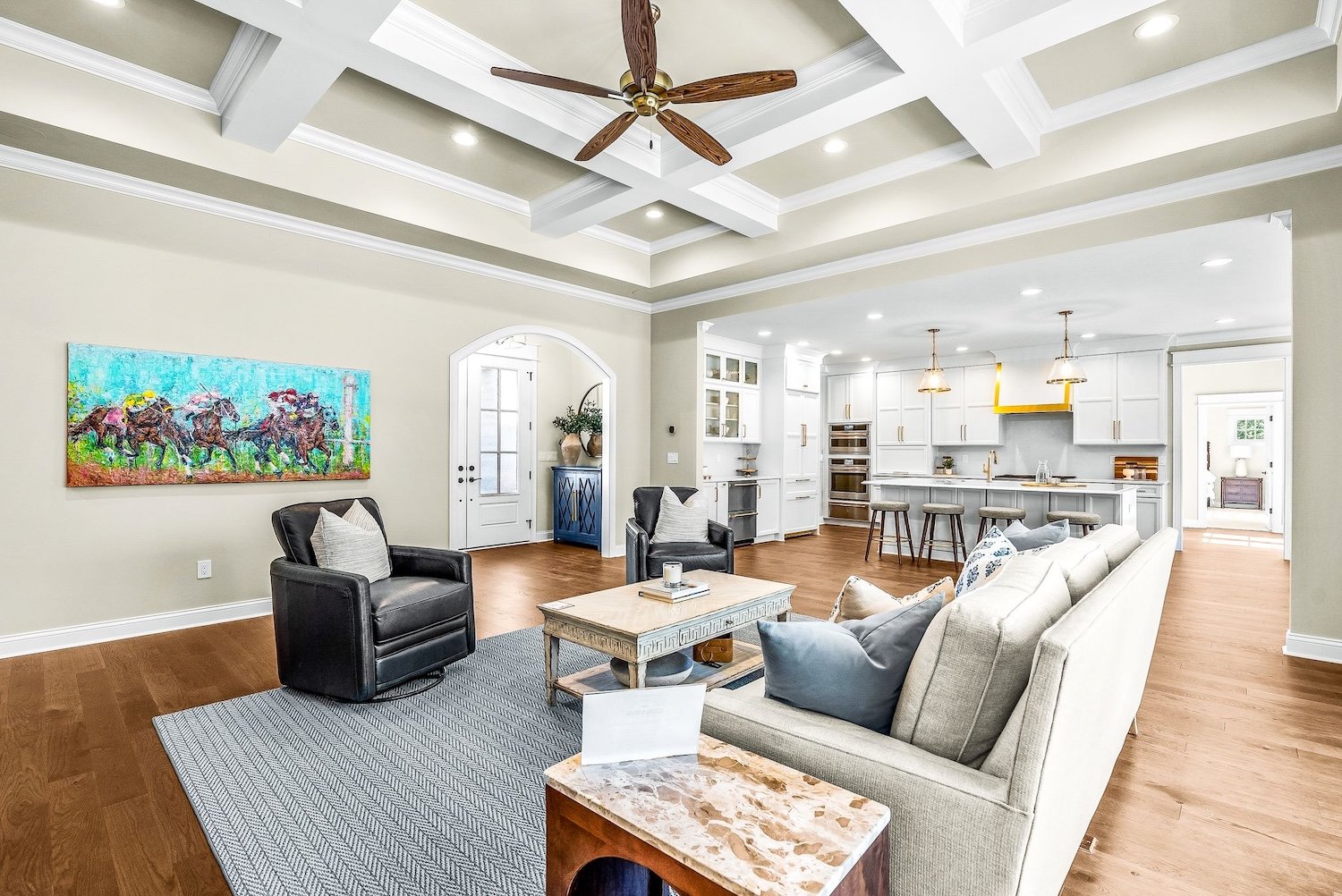 Open-concept living room and kitchen with high ceiling, white walls, hardwood floors, and modern furnishings. A painting of horse race on the wall, ceiling fan, bar stools at kitchen island, and various decorative items.