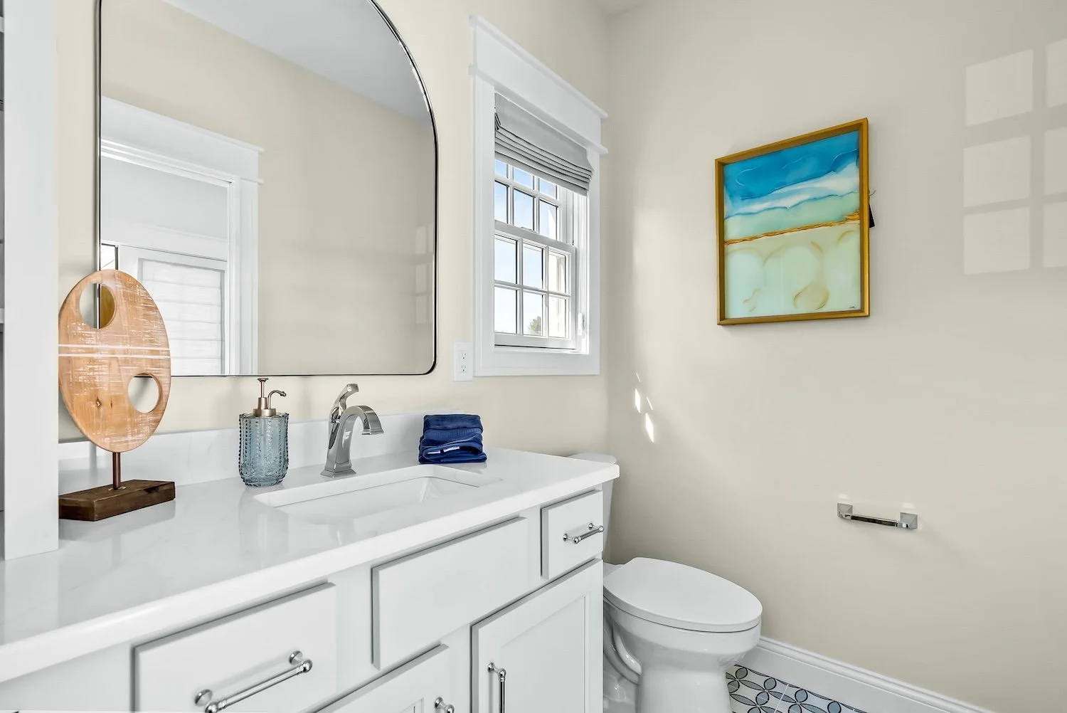 Bathroom with white vanity and sink, large mirror, soap dispenser, folded blue towel, window, landscape painting on beige wall, toilet, patterned floor.