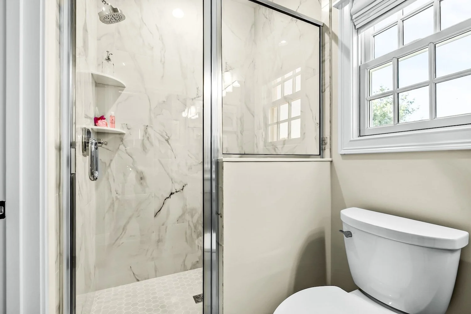 A bathroom with a glass-enclosed shower area with marble walls, a white toilet, and a large window with a white frame, letting in natural light.