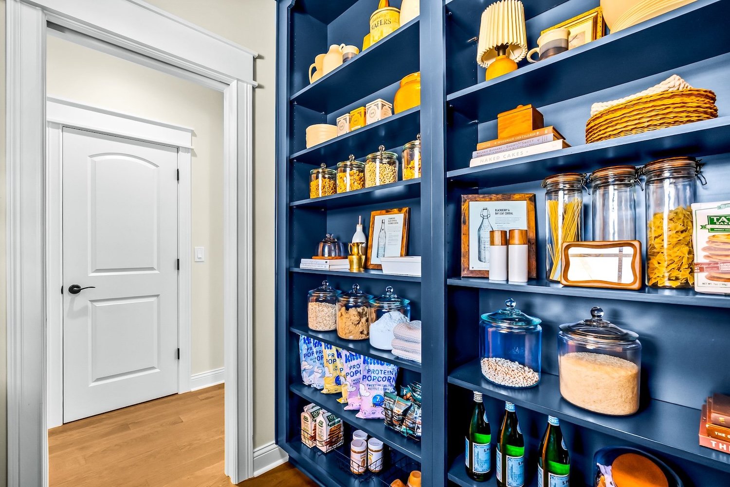 A pantry with dark blue shelves filled with various jars, boxes and bottles, next to a white door with molding and a black handle, on a light-colored wall with wooden flooring.