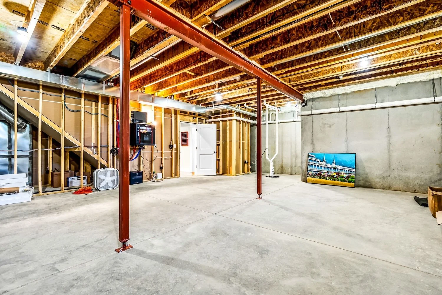 Empty unfinished basement with concrete walls and floor, exposed wooden ceiling beams, metal support post, staircase, small fan, and a large framed picture of a building leaning against the wall.