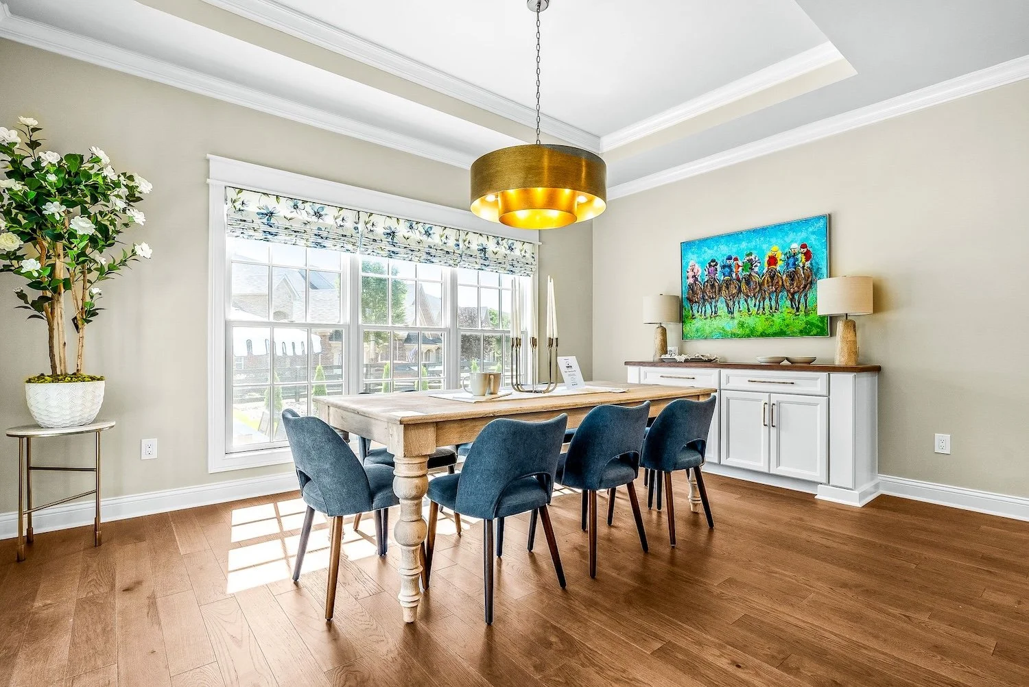 A bright dining room with hardwood floors, a large window with floral valance, a wooden dining table with six blue chairs, a white sideboard with lamps and a colorful painting of a horse race above it, and a potted plant in the corner.