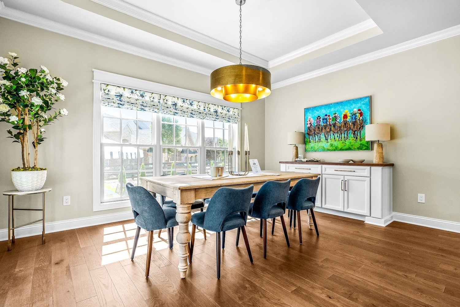 A bright dining room with hardwood floors, a large window with floral valance, a wooden dining table with six blue chairs, a white sideboard with lamps and a colorful painting of a horse race above it, and a potted plant in the corner.