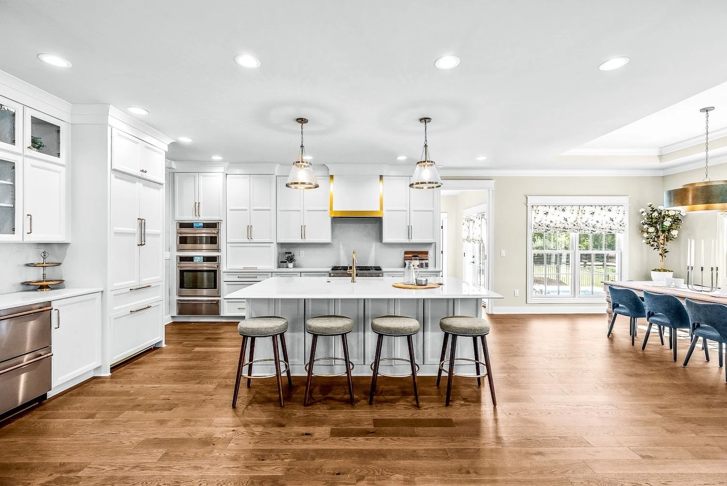 Bright, modern kitchen with white cabinetry, a large island with four stools, hardwood floors, and stainless steel appliances. There are windows and a dining area with blue chairs in the background.
