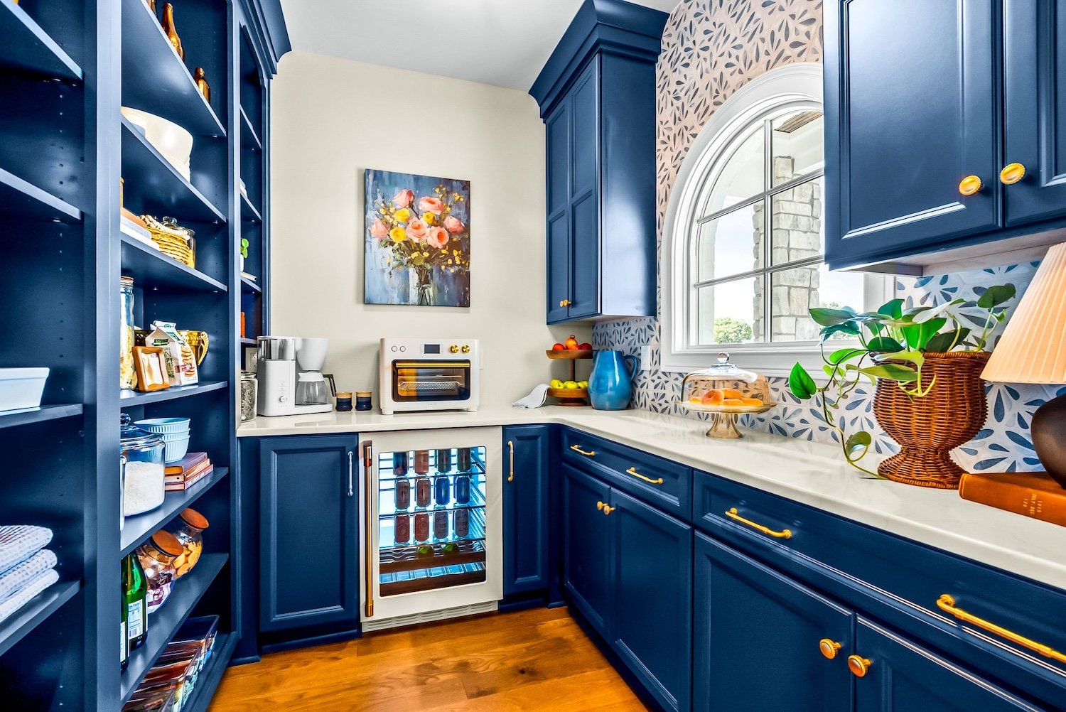 A colorful kitchen with navy blue cabinets, a white countertop, a window with a view outside, and decorative items like a blue pitcher, a cake stand, and a portrait painting on the wall.