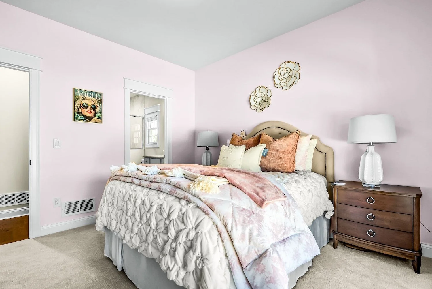A bedroom with pastel pink walls, a queen-sized bed with beige upholstered headboard, decorated with various pillows and a pink throw blanket. There are two white lamps on dark wood nightstands, metal flower wall art above the bed, a Vogue magazine c