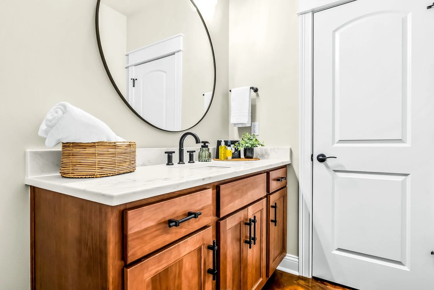 Bathroom vanity with wooden cabinets, a white marble countertop, and a black faucet. A round mirror hangs above the sink, and a wicker basket with rolled white towels is on the counter. Small bottles and a green potted plant also sit on the counter. 