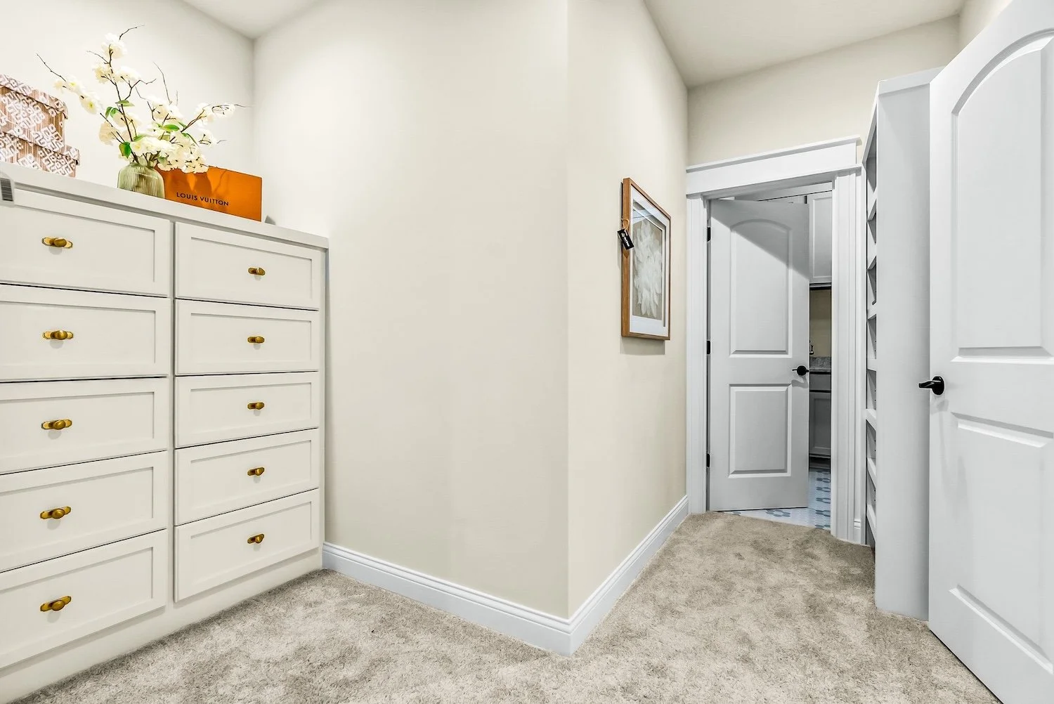 Interior view of a room with a white dresser with gold knobs, a flower vase with white flowers and a Louis Vuitton box on top, a framed picture on the wall, and open doorways leading to another room with visible tile flooring.