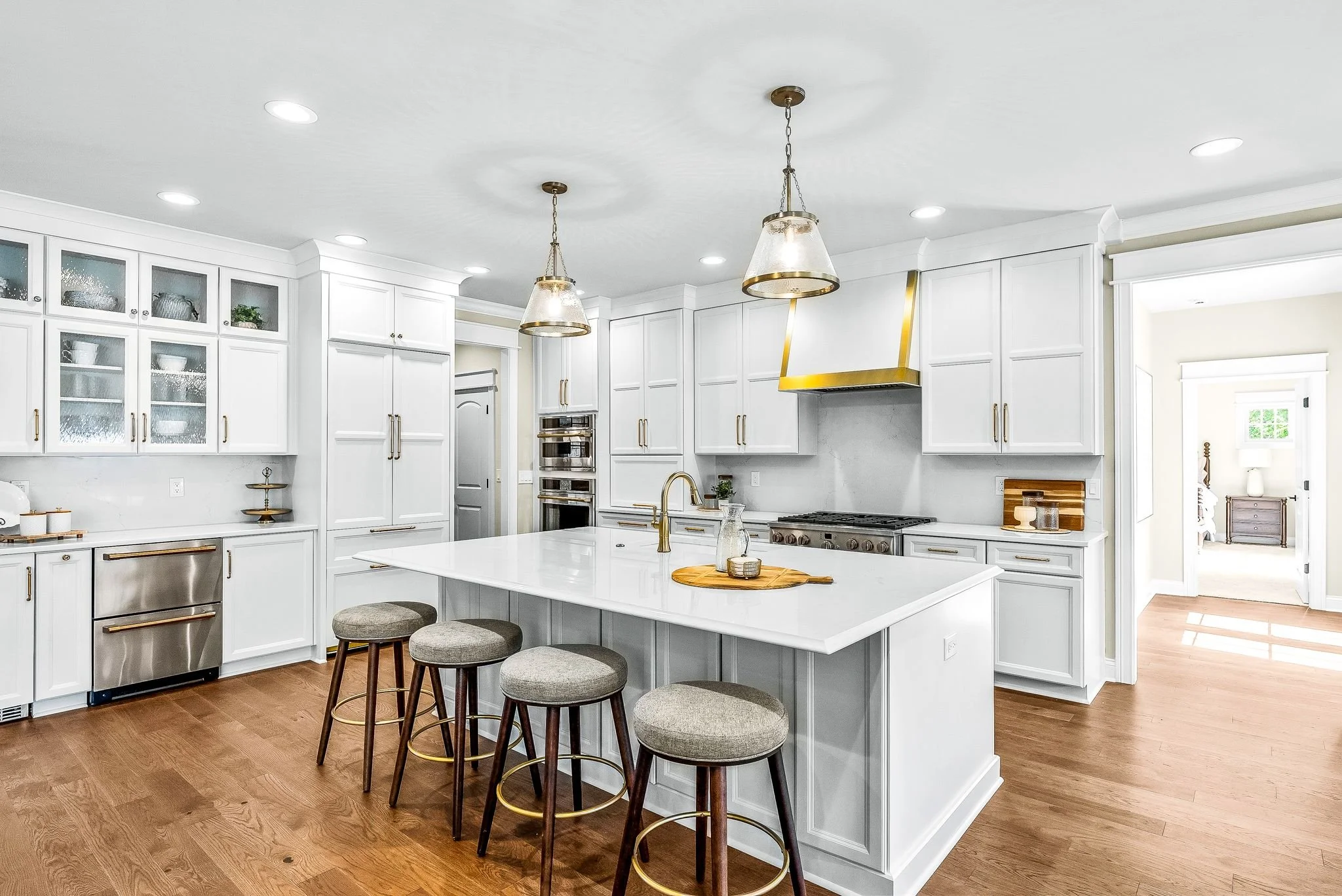 Modern white kitchen with wooden flooring, pendant lights, and a kitchen island with bar stools.