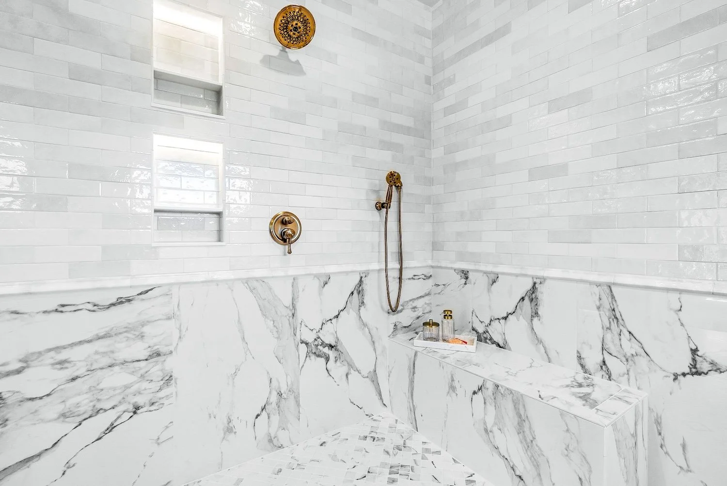 Modern walk-in shower with white subway tile walls, marble wainscoting, a built-in bench, a brass hand shower, and a showerhead. Small built-in niches hold toiletries. There is a window and a decorative wall hanging.