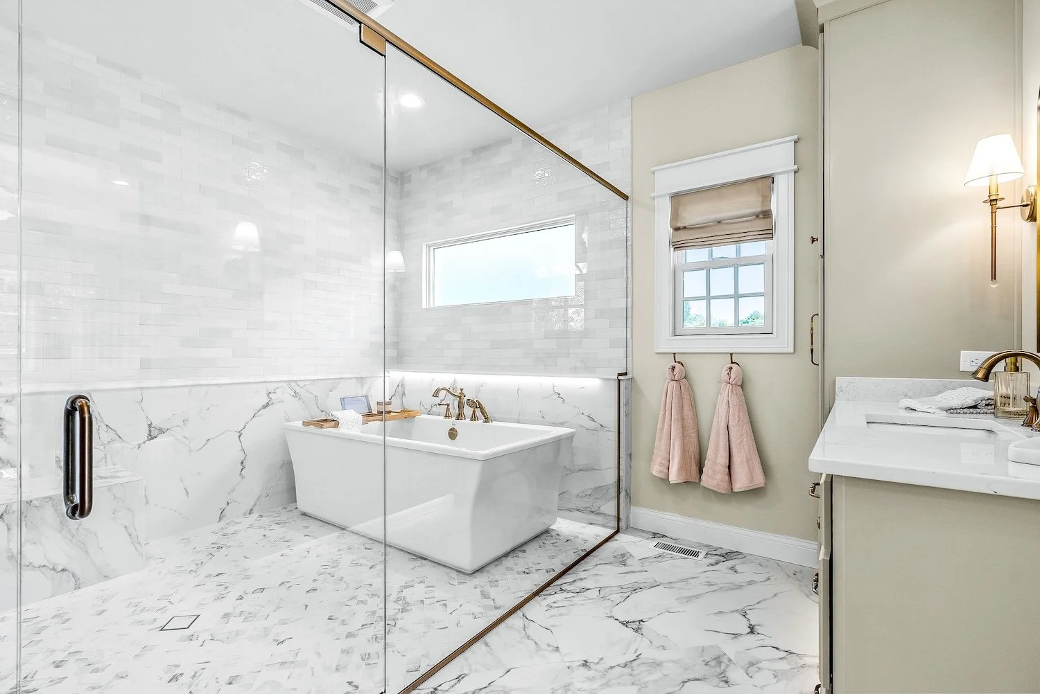 Bright bathroom with a white marble floor and wall tiles, a frosted glass shower enclosure, a standalone bathtub with gold fixtures, a small window, and a bathroom sink with a marble countertop.
