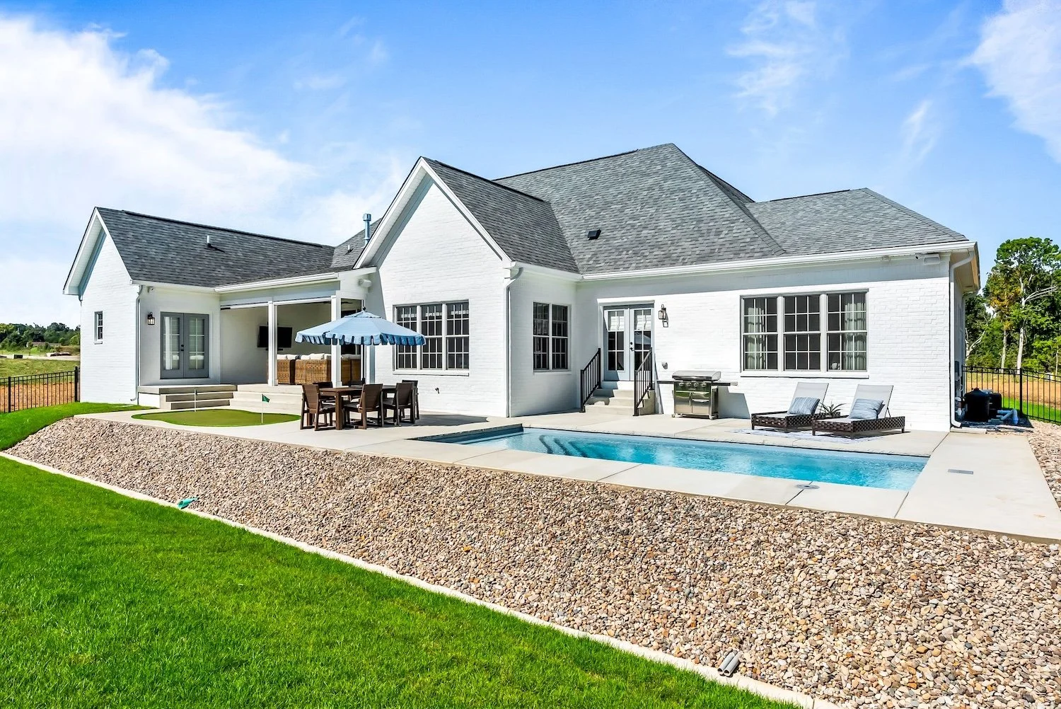 White modern house with a backyard pool, outdoor dining area with umbrella, lounge chairs, and well-kept green lawn under a partly cloudy sky.