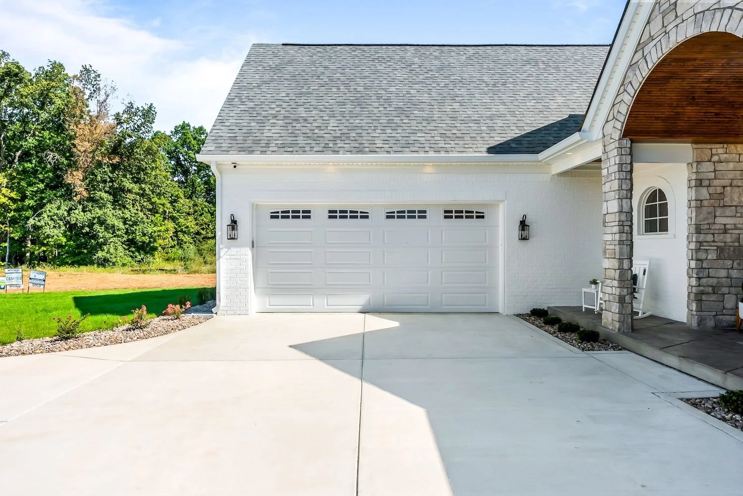 Front view of a white garage door attached to a house with a gray roof, stone and white exterior walls, and a small porch with outdoor furniture.