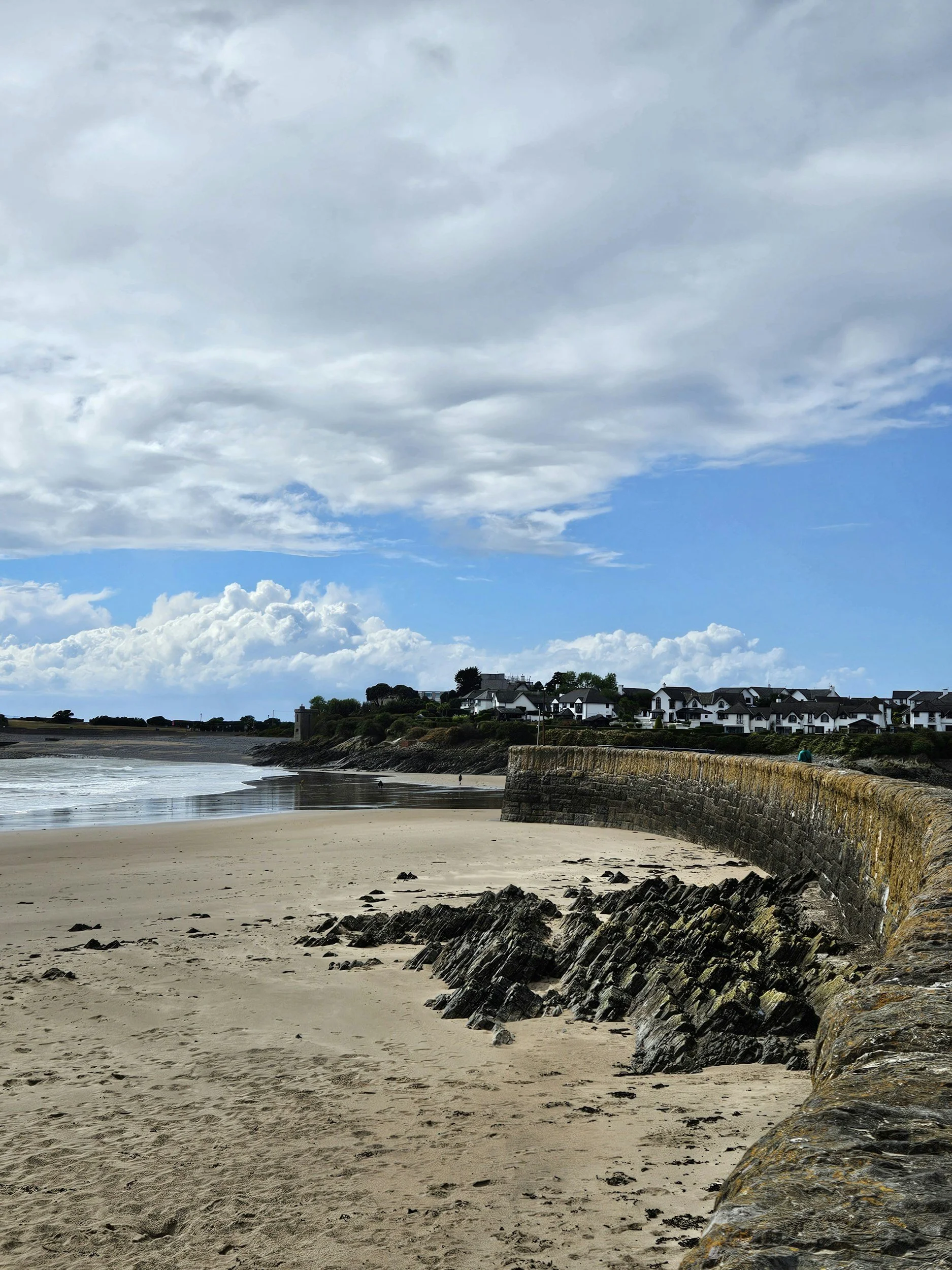 A beach with sandy shore, rocky area, stone wall, and waves in the background under a partly cloudy sky.