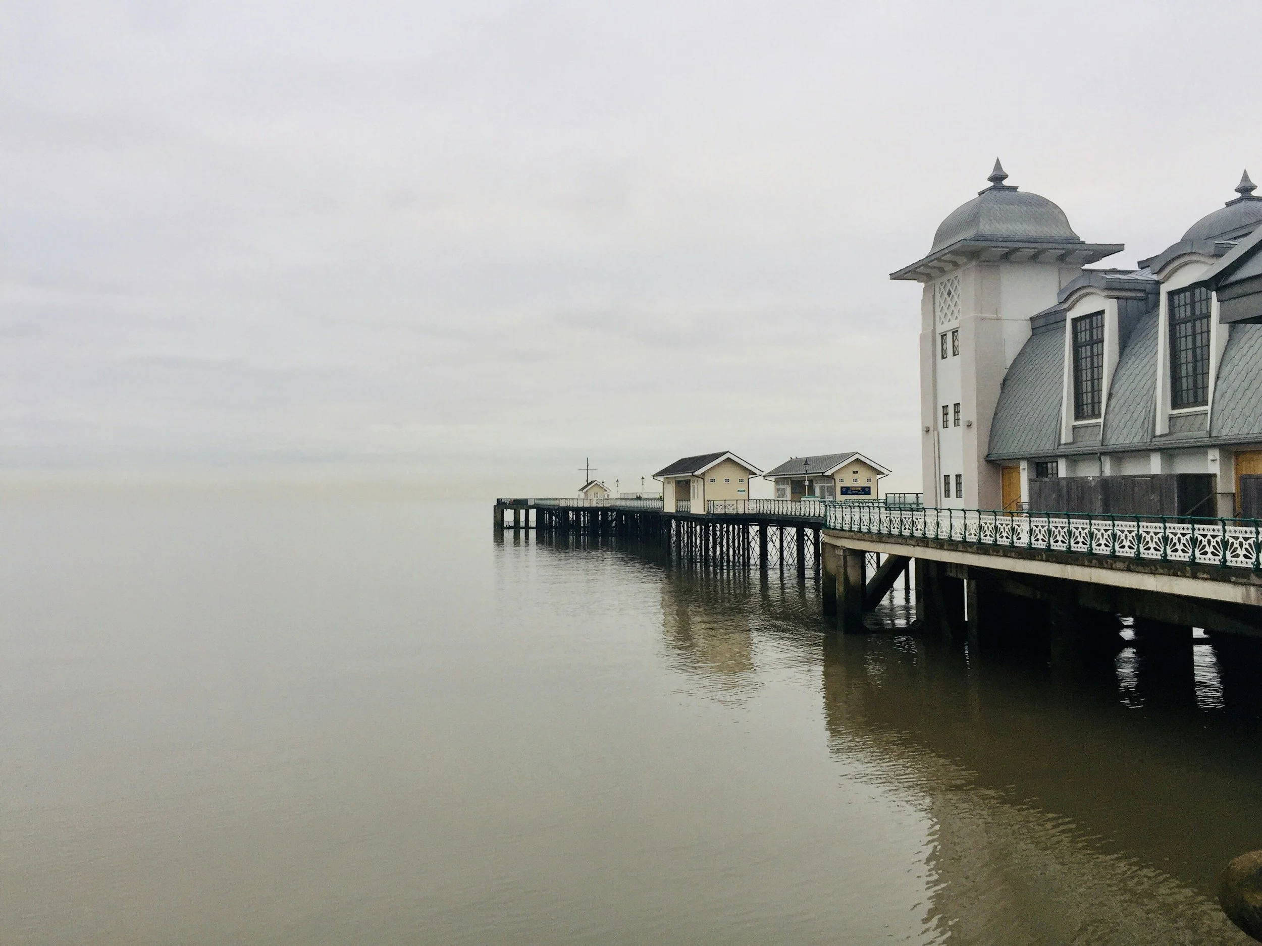A pier extending into the water with houses and a building that has a steeple, in a calm, overcast setting.
