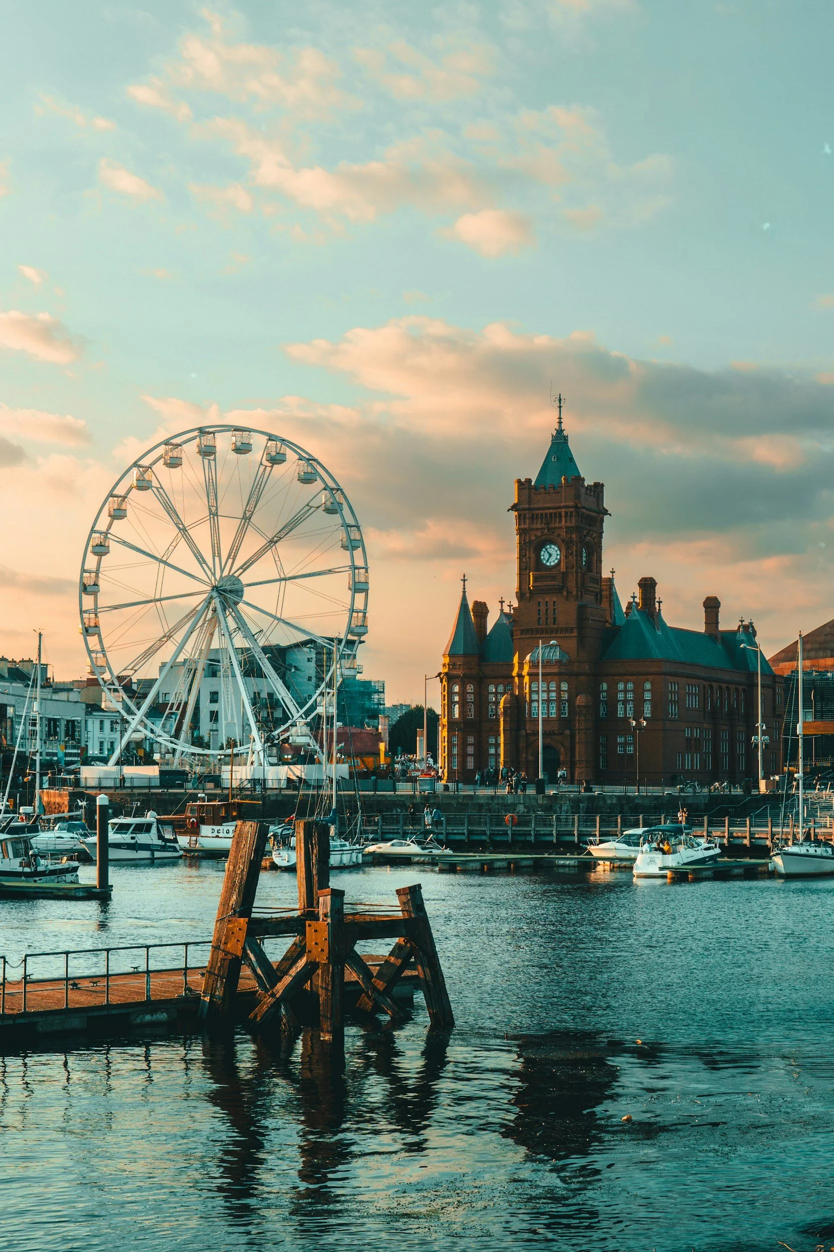 A cityscape featuring a large Ferris wheel, a historic clock tower building, and a marina with boats and water reflections in the foreground during sunset.