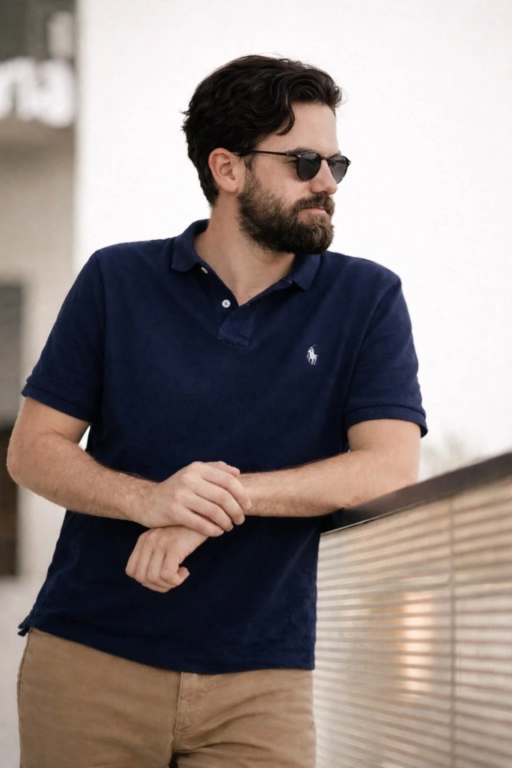 A man with dark hair, a beard, and sunglasses leaning on a railing, wearing a navy blue polo shirt and tan pants.