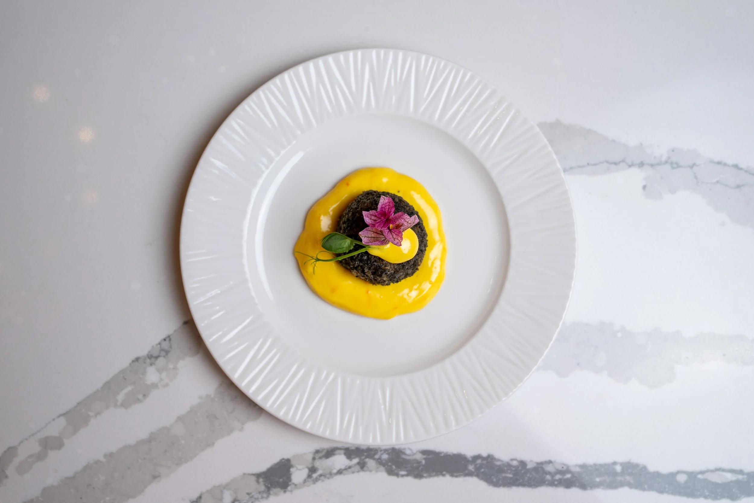 A plate with a fried black dumpling topped with pink edible flower, green leaves, and yellow sauce, on a white textured plate on a white marble surface.