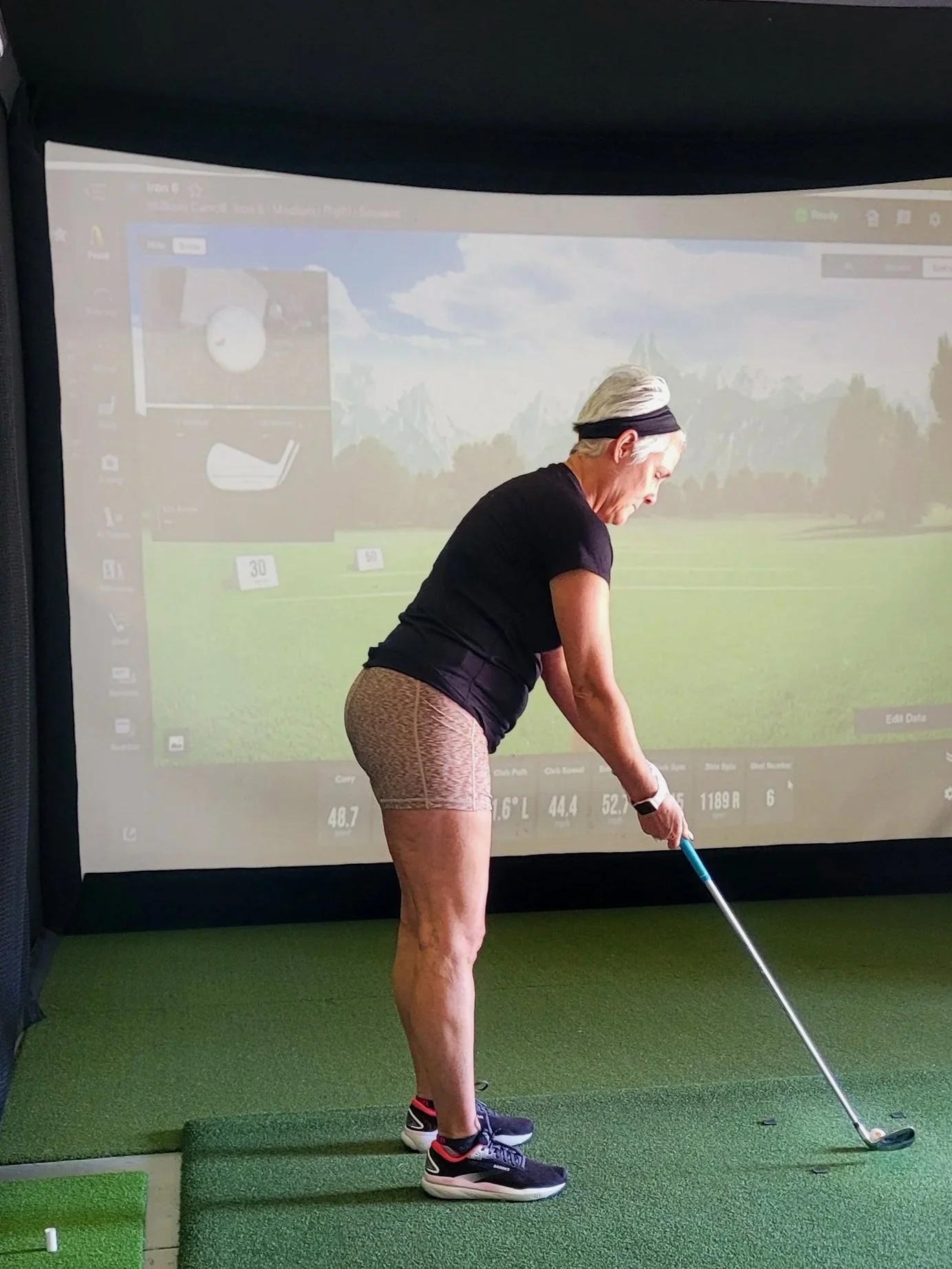 Older woman in black shirt and beige shorts practicing golf swing indoors on green turf with a virtual golf simulator in the background.