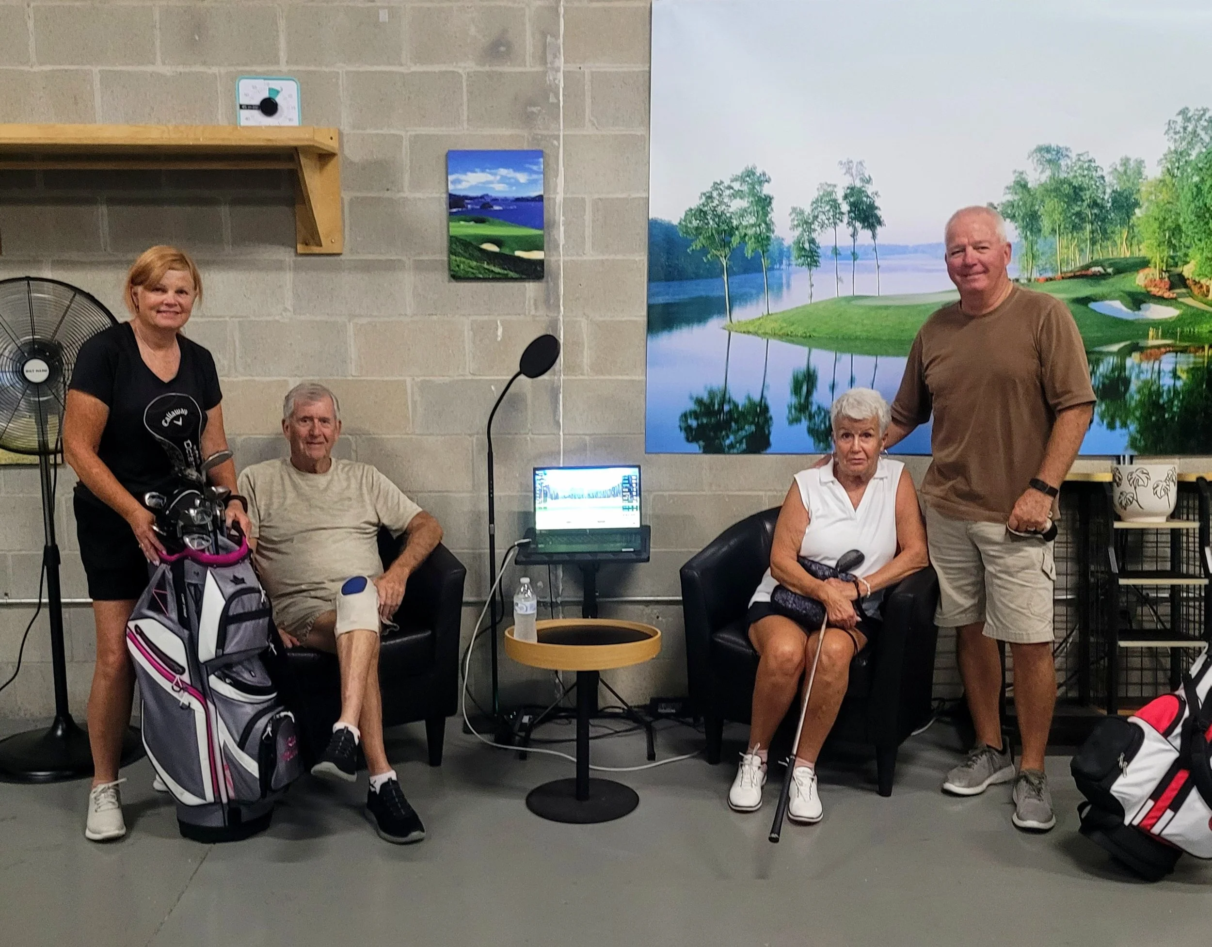 Four elderly people in a golf-themed indoor setting, two seated in black chairs and two standing. A woman with a golf bag and a woman with a golf club are seated, while a man and another woman stand nearby. A large landscape painting of a golf course is on the wall behind them, and a small table with a water bottle and laptop is between the seated individuals.