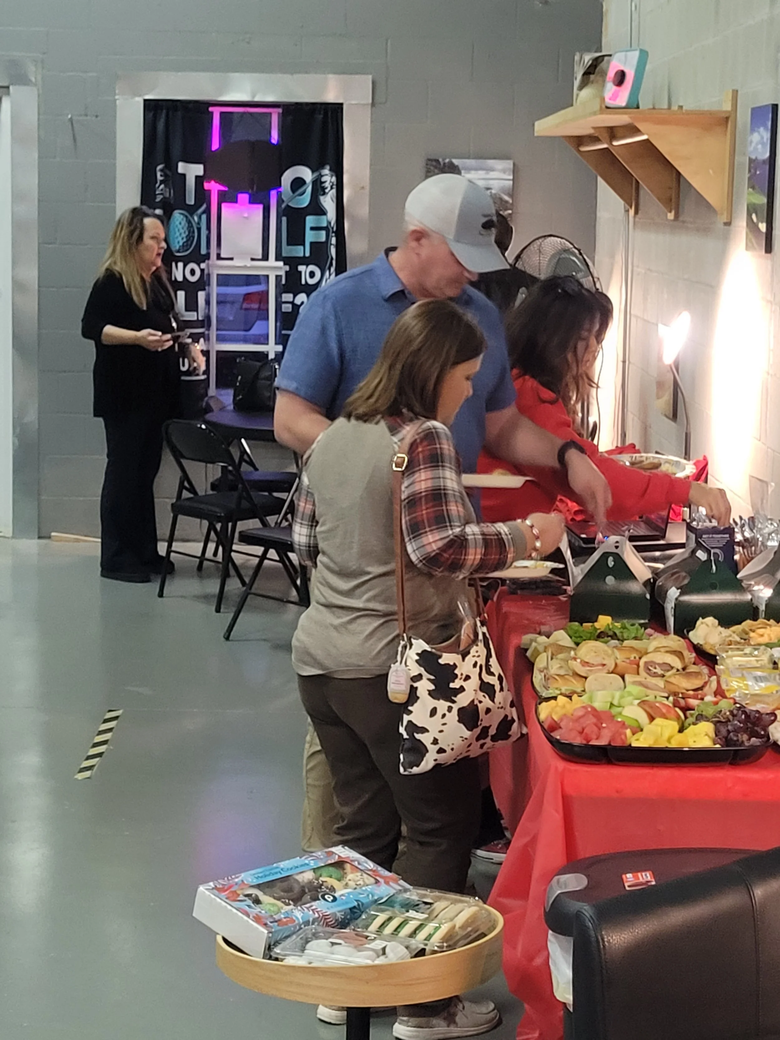 People at a buffet table with fruit, sandwiches, and snacks, in a room decorated for a social gathering.