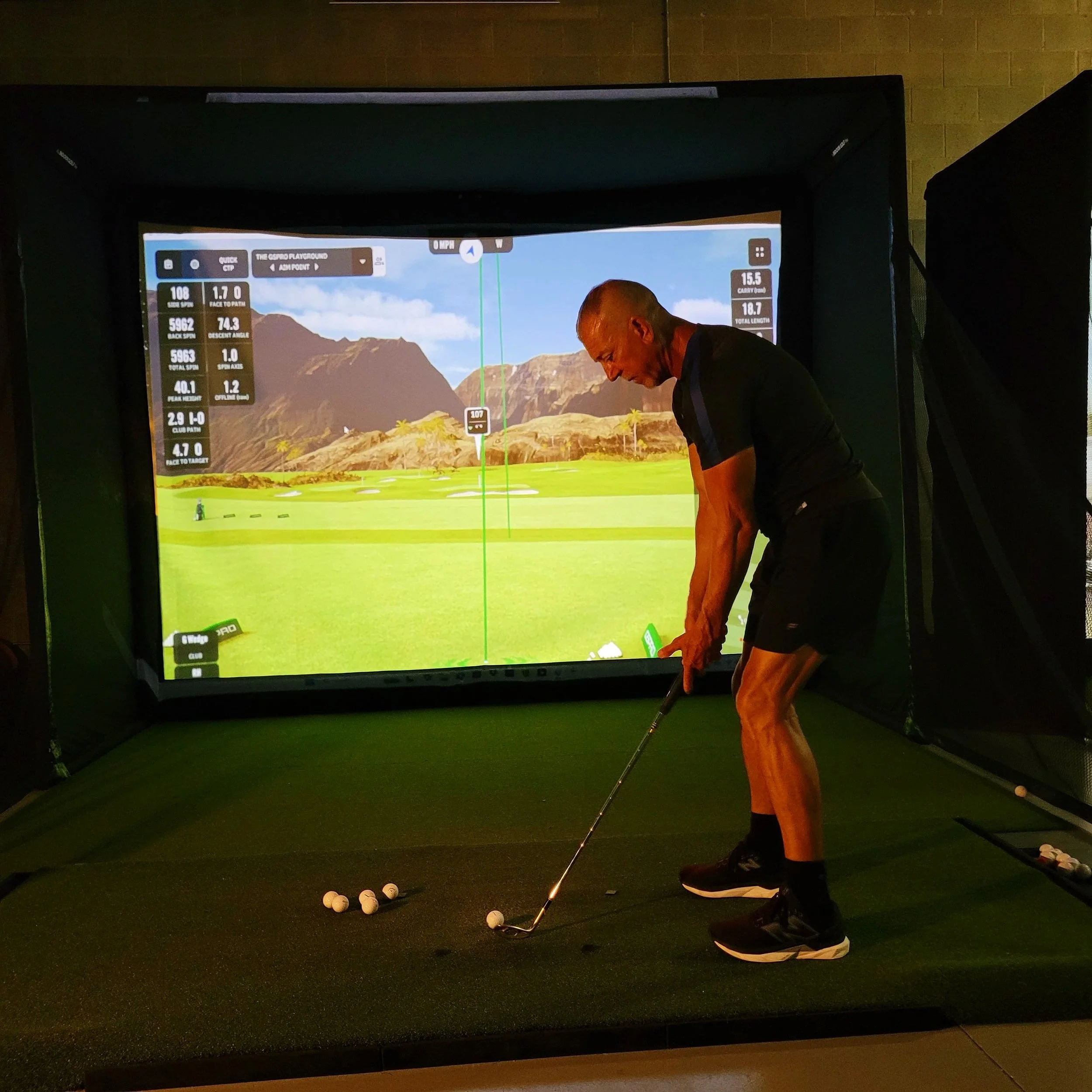 A man practicing golf indoors on a golf simulator with a screen showing a virtual golf course in the background.