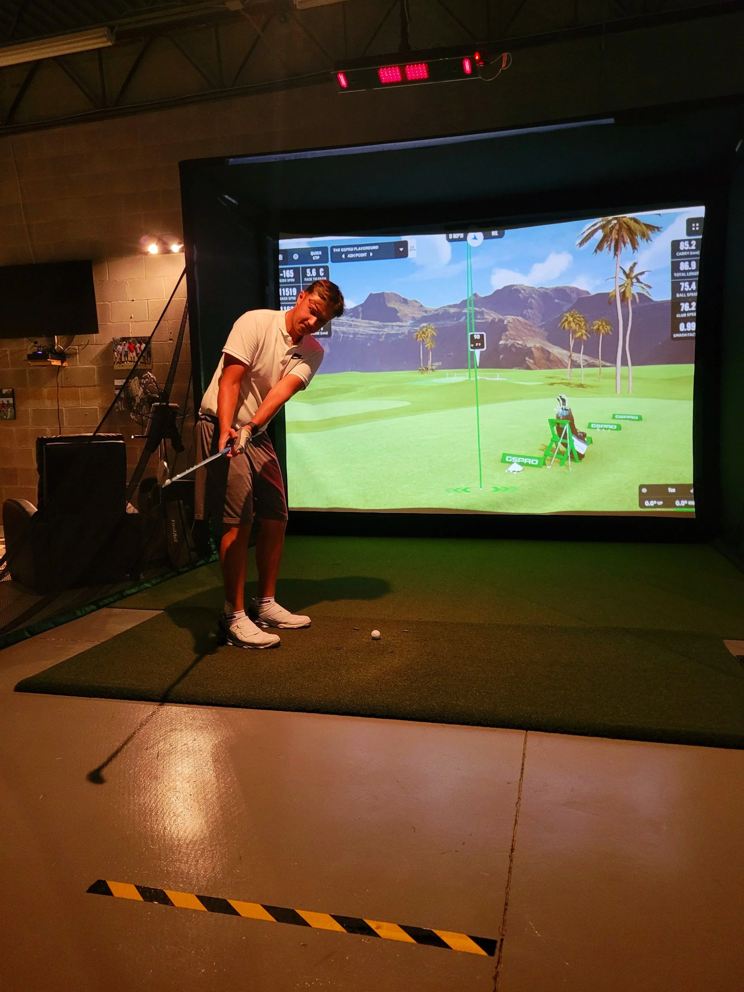 A person practicing golf indoors using a golf simulator. The person is holding a golf club and standing on a small green mat, with a large screen displaying a virtual golf course in the background.