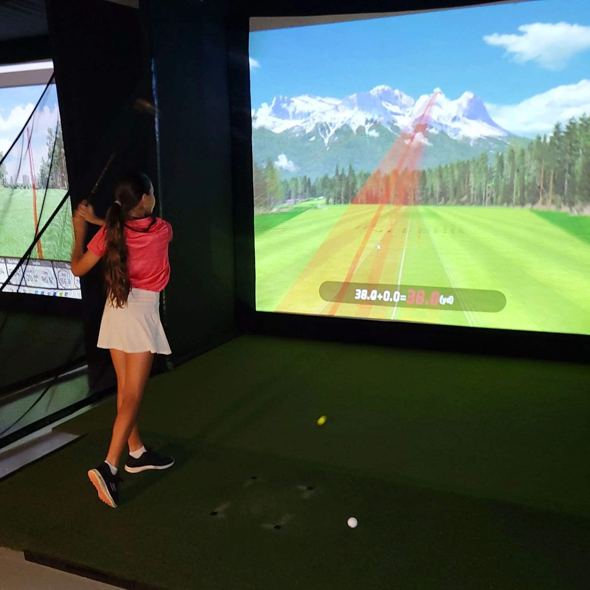A girl playing indoor golf on a simulator, swinging a golf club while looking at a large screen displaying a scenic mountain landscape and a golf course.