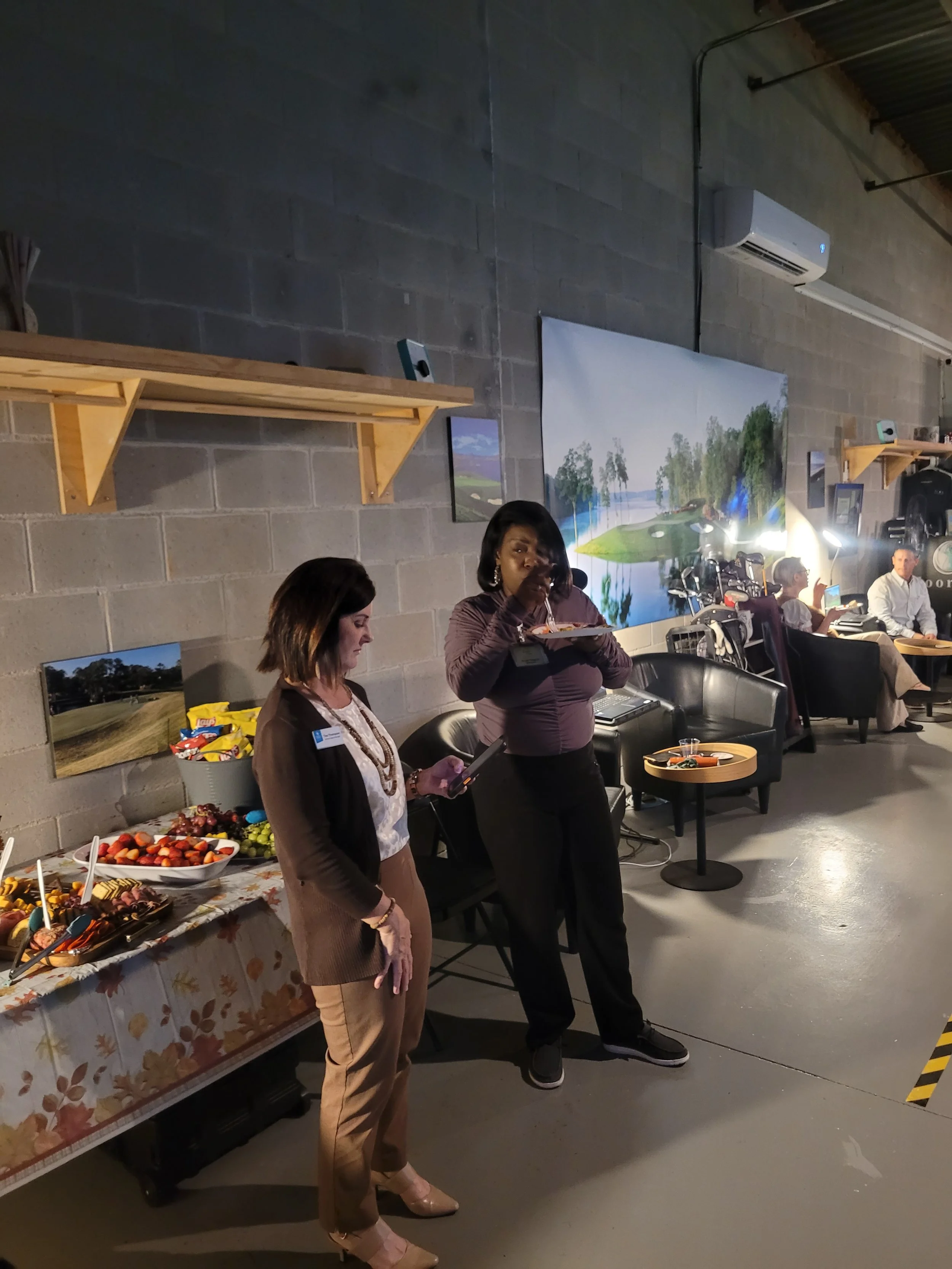 Women standing near a buffet table with snacks and strawberries in a conference or event space, with some people sitting and chatting in the background.