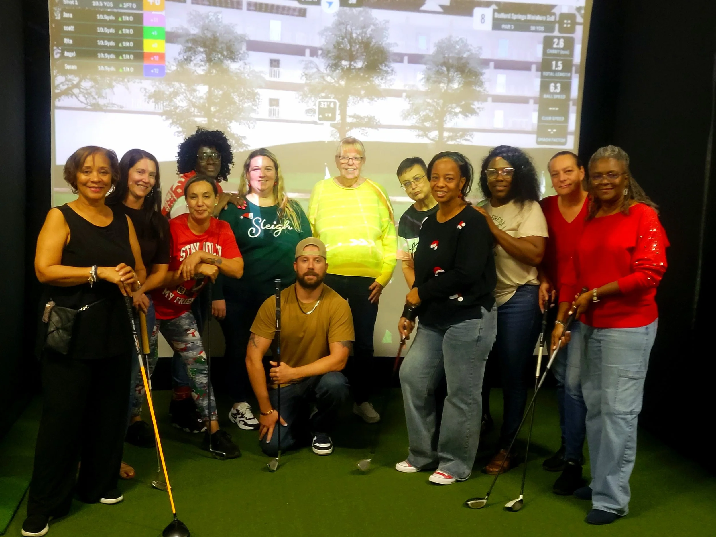 A group of 12 diverse women and one man posing indoors in front of a large screen displaying golf scores and stats. Some women are holding golf clubs, and the man is kneeling with a golf club. The women are dressed casually, with some wearing festive or sporty attire.