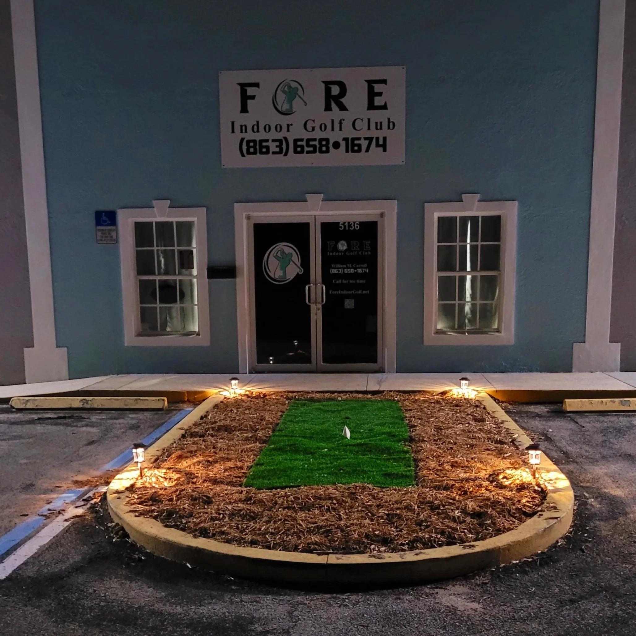 Exterior view of a building with a sign reading 'FORE Indoor Golf Club' with phone number, and a small entrance with double glass doors. There is a small landscaped area with artificial grass and lights in front of the entrance, surrounded by parking spaces.