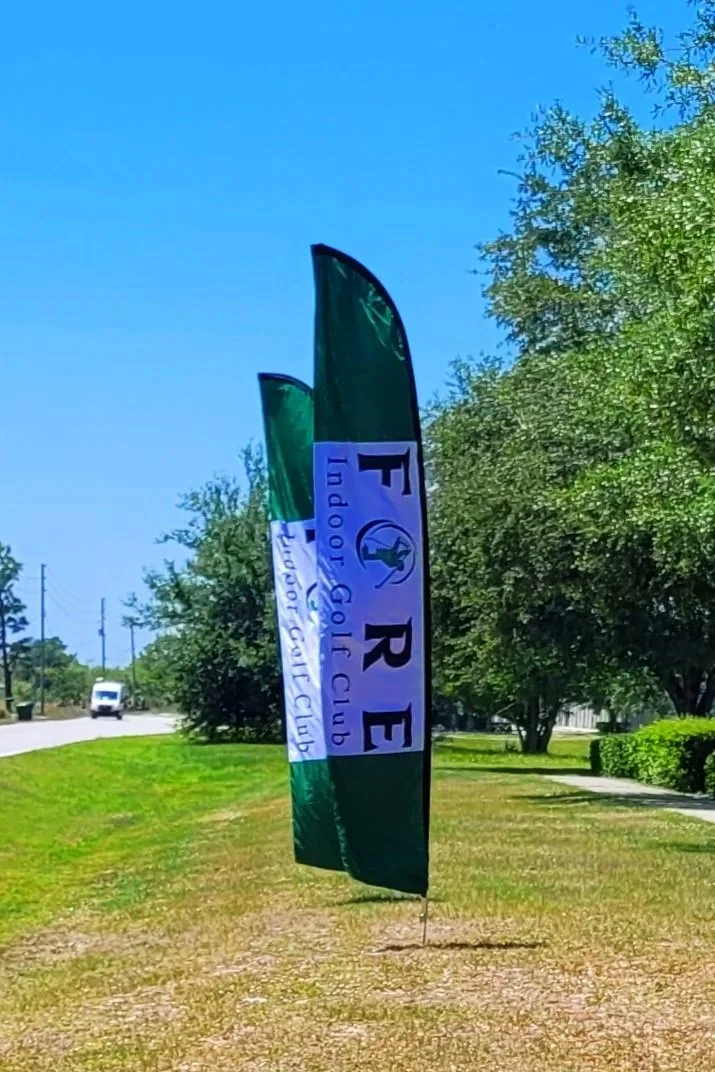 A green and white feather-shaped flag for the Ford Golf Club stands on a grassy area near a sidewalk, with trees, bushes, and a street with a vehicle in the background under a bright blue sky.