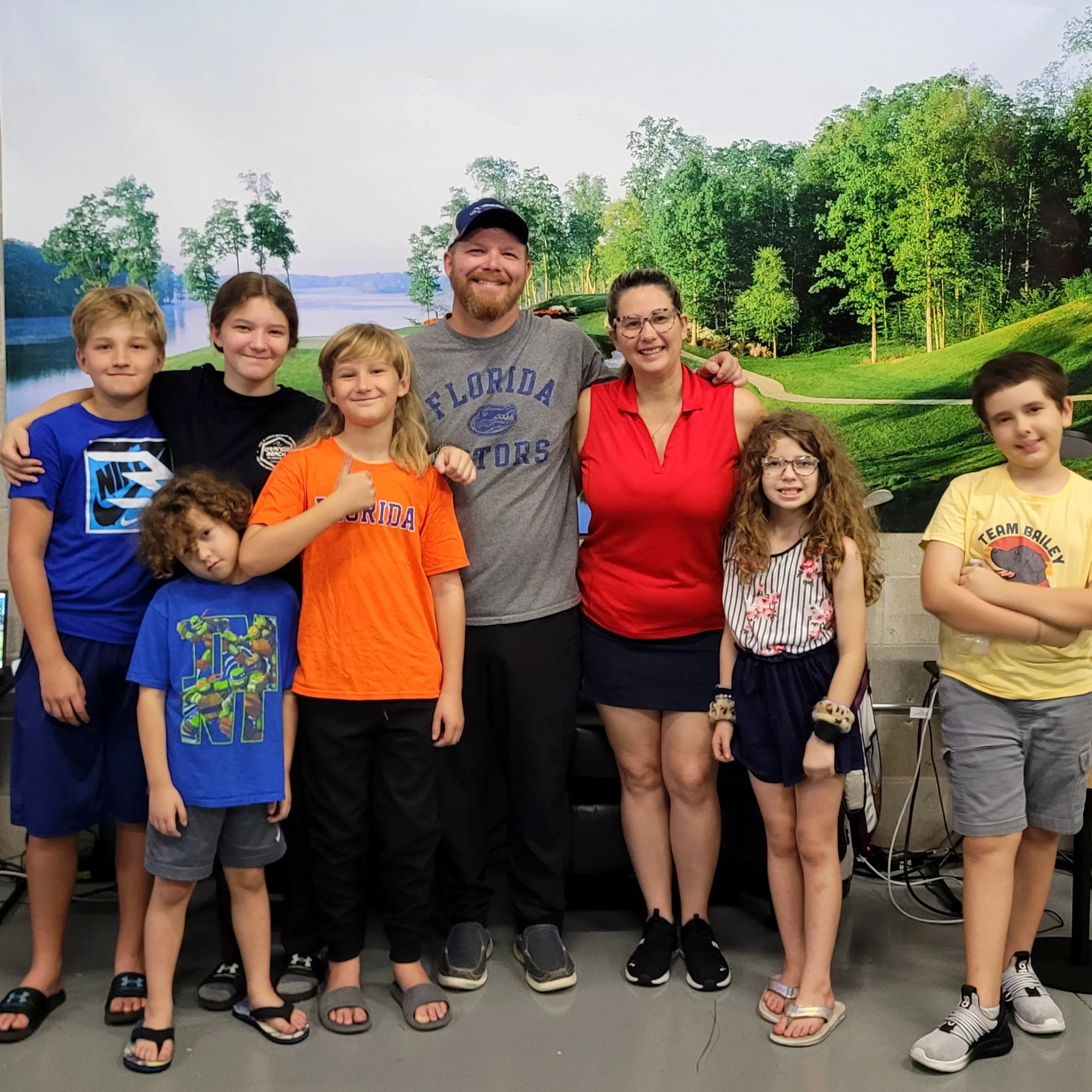 Family group of nine people, three adults and six children, smiling and posing together indoors in front of a scenic landscape mural.