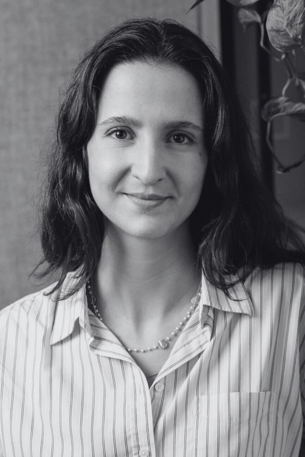 Ines, a woman with wavy, shoulder-length hair, wearing a striped button-up shirt, smiling in an indoor setting with glass and metal structures in the background.