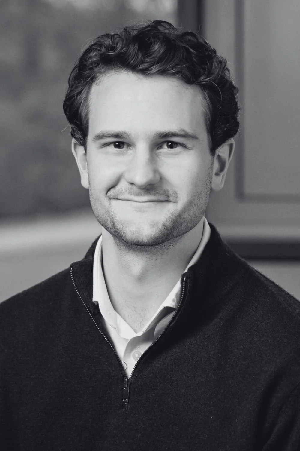 Black and white portrait of Sven, a young man with short hair, light beard, and a slight smile, wearing a Patagonia fleece jacket and a collared shirt, with bookshelves in the background.