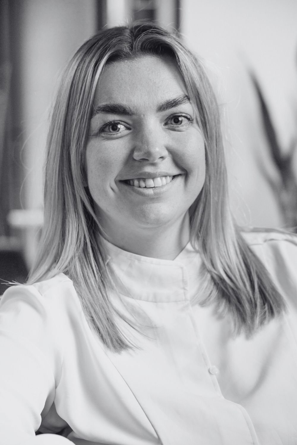 Bethany, a woman smiling with straight hair sitting indoors, with large windows and blurred background.