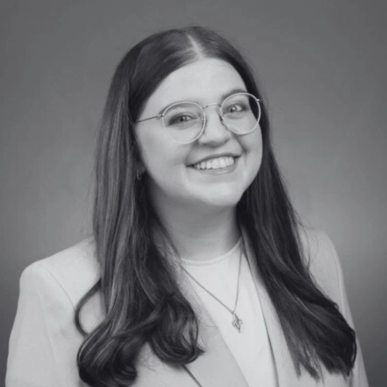 Black and white photograph of Kelsey, a young woman with long, wavy hair, wearing glasses, a blazer, and a necklace, smiling at the camera against a plain background.